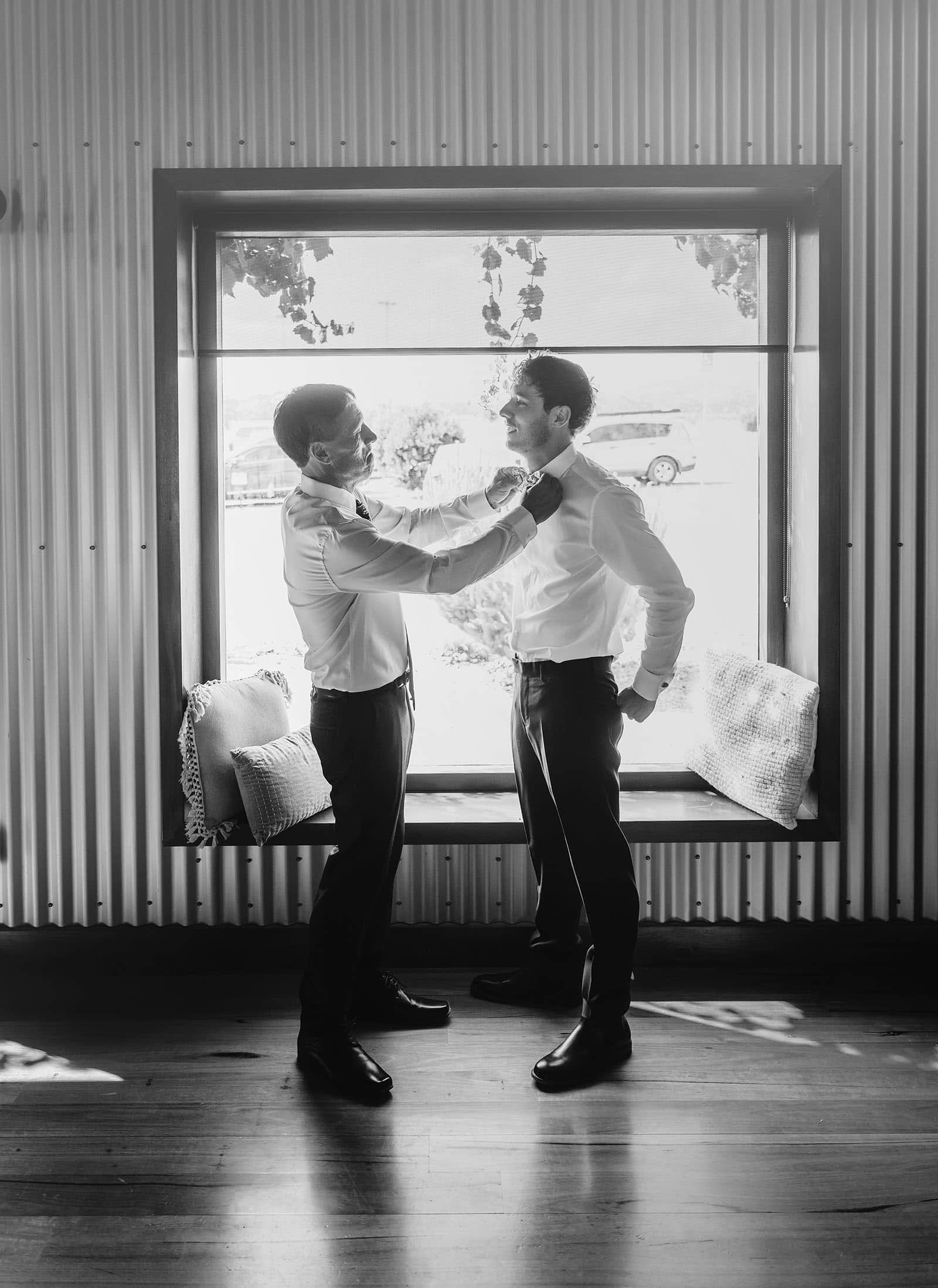 tender bond: two men in formal dress by window Two men in formal attire share a tender moment by a large window.